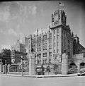 A towering, fenced in building with a clock tower and flagpole on one corner, in black-and-white.
