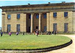 A large stone built building of two stories. In the middle are two columns and on both sides two tall windows on the ground floor, and two smaller windows on the first floor. In the foreground a grassed area; and just visible a circular gravelled area leading up to the front of the building.