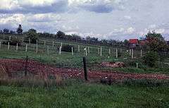 A hillside with multiple barbed-wire fences running parallel to each other, with fruit trees, a barn and a watchtower in the background.
