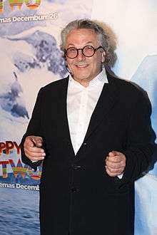 A male with grey hair and round black-rimmed glasses is seen standing in front of a blue and white wall. He is wearing a black jacket on top of a white open collared shirt and is smiling.