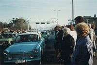 East German Trabant cars driving between dense crowds of people. Metal gantries over the road and a border watchtower are visible in the background.