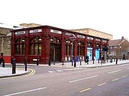Single storey red glazed terracotta station building with tall arched windows. Glass shaded lamps project from the façade on ornate brackets and the words "Kilburn Park", "Entrance", "Exit" and "Underground" are displayed in black on a white moulded tile band above the window arches.