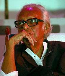 A photograph of an old man wearing glasses and right-hand kept on his chin