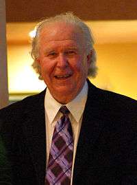 A man with disheveled white hair wearing a black suit jacket and purple tie looks straight ahead and smiles.