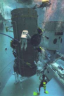 Underwater photograph of the Neutral Buoyancy Space Simulator, showing divers and a full-scale model of a spacecraft in the tank.