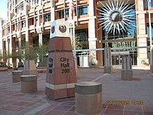 photos showing the short obelisk signage showing City Hall, and topped with the seal of the city, a stylized maroon phoenix. The semi-circular front of the building in the background, adorned with a stylized sunburst.