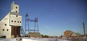 alt=A mine shafthouse and hoisthouse in a snowy landscape. A large brick building can be seen in the background.