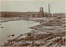Sepia-tone photo of a pond surrounded by large logs. At the far end of the pond is a large building with a square tower and two smokestacks. Label is "T&T L CO MILL POND RICKETTS PA 1903" (i.e. Trexler and Turrell Lumber Company Mill Pond ...)