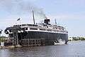 A large black ship with white upper decks rests at a berth, its rear surrounded by wooden posts.  The rear access to its vehicle decks is opened upward.