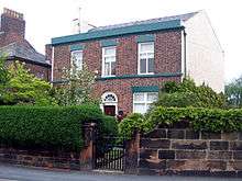 A sandstone wall with a hedge and a gate in front of a plain brick house in two storeys with three bays and an arched central doorway; the right side of the house is painted white and the edge of a larger house is to the left