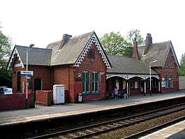 Two sets of rails between platforms beyond which is a brick building with two gables and a canopied area between them; a sign to the left says "Widnes"